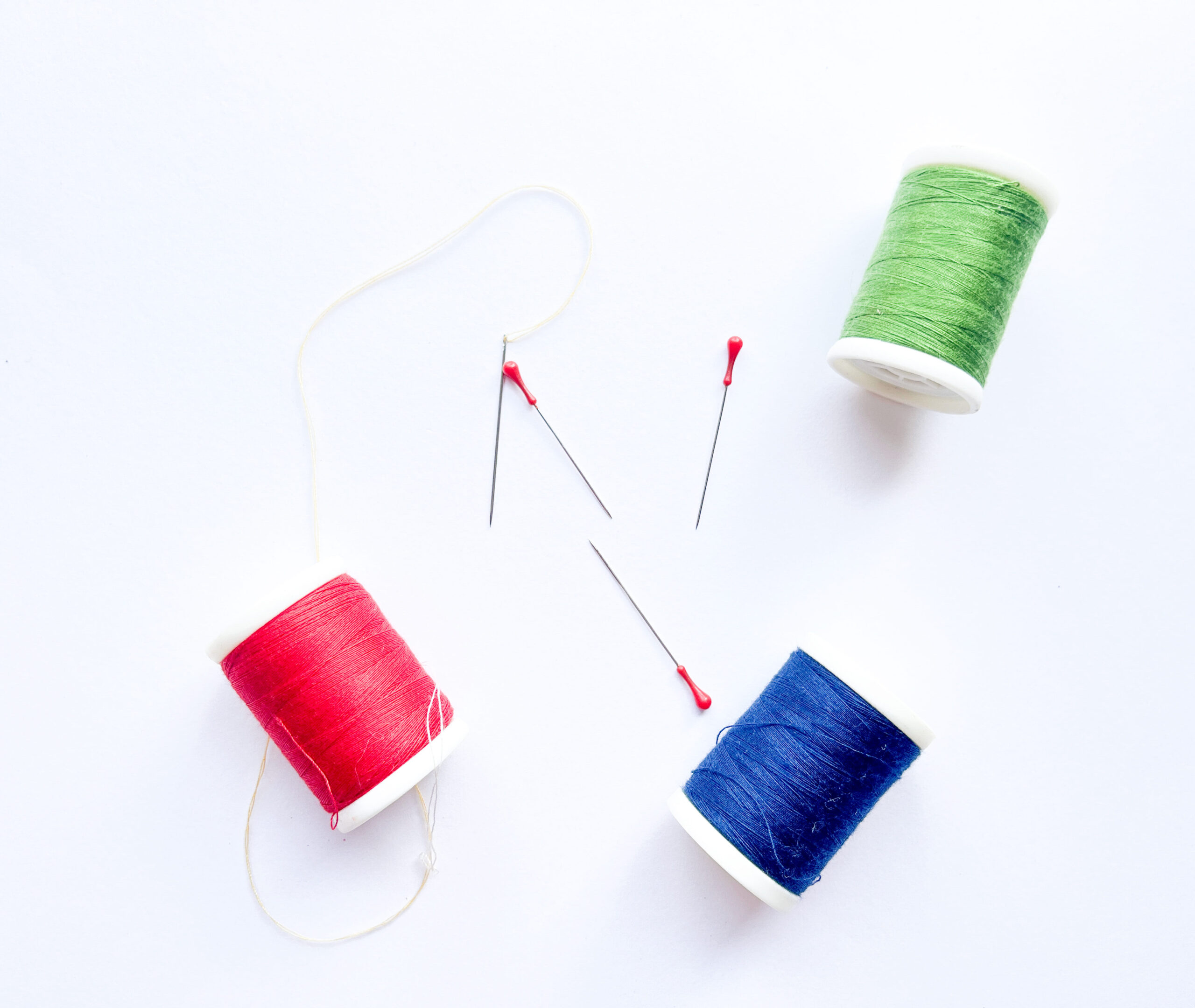 three spools of thread of red, blue and green colors with needls on white surface view from top